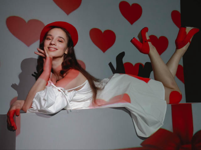 StacieLove with red hat, white dress, and red heels, lying on her stomach, smiling, hands on cheek, legs up, showing her ass, hearts on background, in a photo.