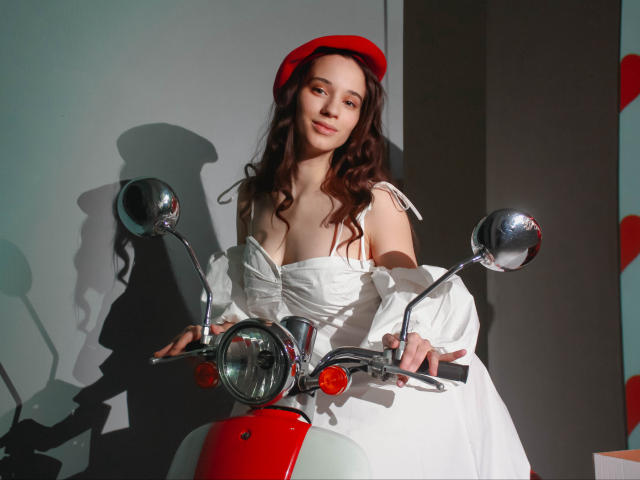 StacieLove, com cabelo castanho ondulado, boina vermelha, blusa branca com um ombro descoberto, e uma scooter vermelha, segurando o guidão, sorrindo, reflexo nos espelhos, sombra na parede, luz forte..