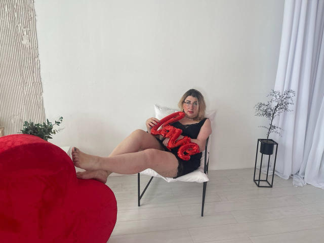 HelenEnigma, a piedi nudi, con un abito nero, che tiene un cuscino rosso con la scritta "AMORE", gambe incrociate, seduta su una sedia bianca, una sedia a forma di cuore rossa in primo piano, pavimento bianco, piccolo supporto nero per piante..