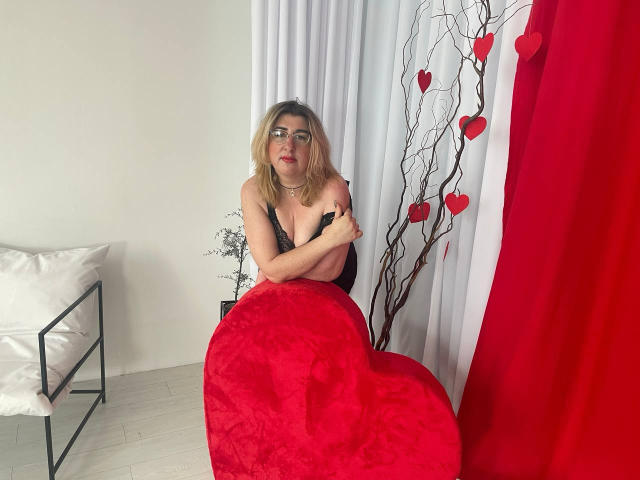 HelenEnigma, avec des cheveux blonds, des lunettes, du rouge à lèvres rouge, un haut en dentelle noire, et une couverture rouge en forme de cœur, les bras croisés, assise sur un sol blanc, avec un coussin blanc, un rideau rouge, et des décorations en forme de cœur..