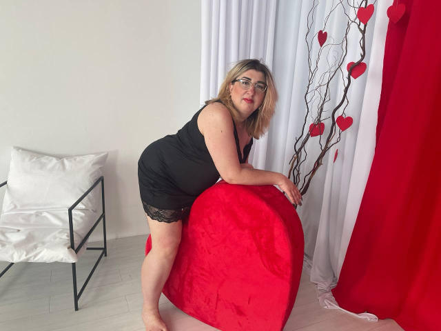 HelenEnigma in a black dress with lace, leaning on a red heart-shaped pillow, showing her ass, with red heart decorations and white curtains in the background.