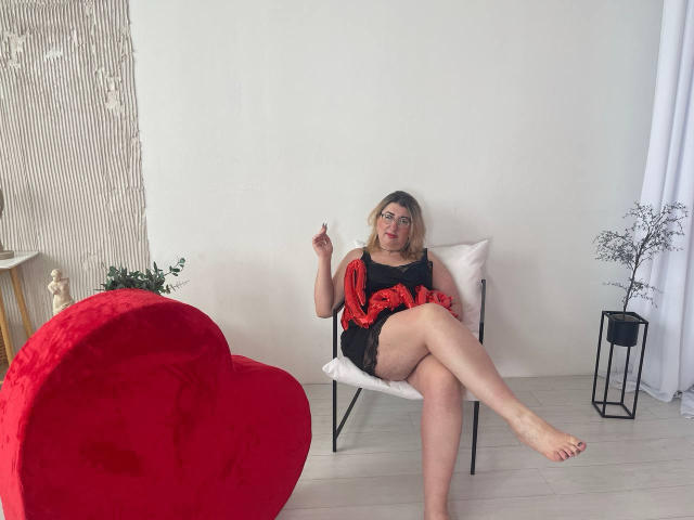 HelenEnigma, blonde avec du rouge à lèvres, portant une robe noire en dentelle avec un motif rouge, assise sur une chaise blanche, une jambe croisée, une chaise en forme de cœur rouge à gauche, un mur blanc, une petite plante..