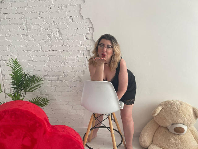HelenEnigma with blonde hair, glasses, black lace dress, leaning on white chair, sucking thumb, red heart pillow, large teddy bear, and green plant in white brick room.