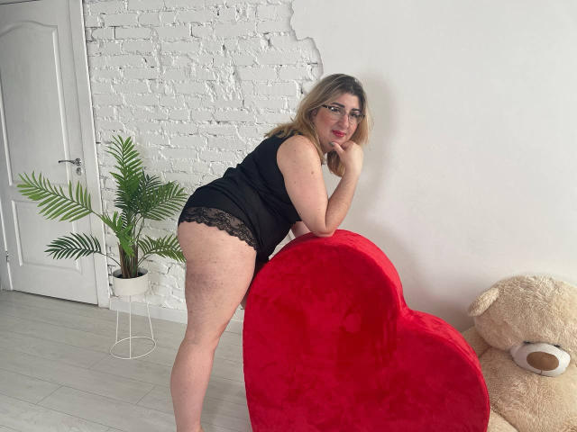 HelenEnigma met een bril, zwarte onderbroek met kantrand, en een zwarte top, leunend op een rood, hartvormig kussen, waarbij haar achterwerk zichtbaar is, blond haar, een witte bakstenen muur, een potplant en een teddybeer..