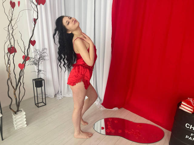 LiyaFrench stands barefoot, wearing red lace lingerie, with long black hair, her hands on her chest, and a red curtain behind her. Her ass is visible.