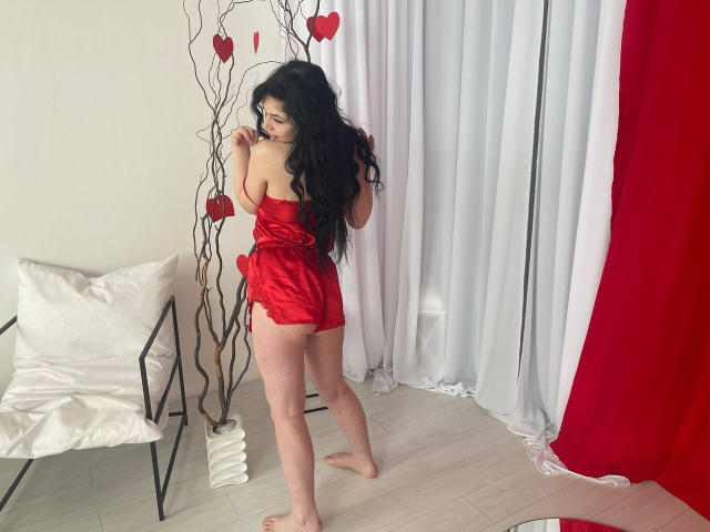 LiyaFrench with long black hair, wearing a red satin romper, showing her ass, standing barefoot, looking back, holding a branch with red hearts. White chair, red curtains.