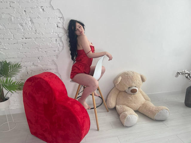 LiyaFrench with long black hair, red velvet dress, sitting on a chair, next to a giant teddy bear and a red heart-shaped couch, barefoot, in a white-walled room.