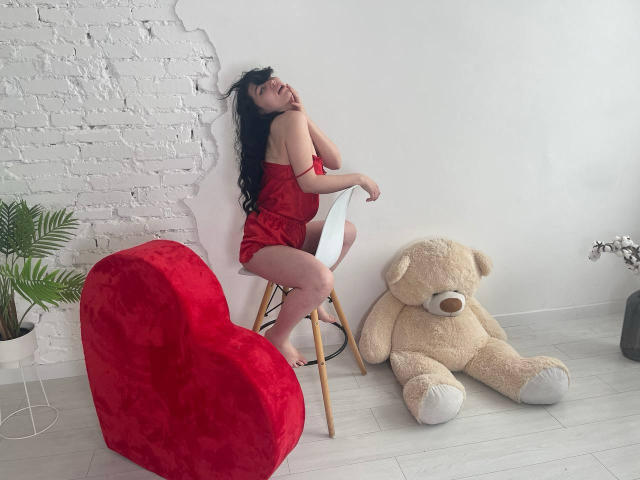 LiyaFrench in red lace dress, sitting on white chair, back against white brick wall, next to giant teddy bear and red heart-shaped pillow, legs slightly apart, black hair.
