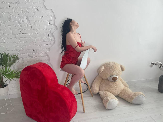 LiyaFrench with long black hair, wearing a red satin dress, sits on a white chair, back against white brick wall, next to a giant teddy bear and red heart chair.