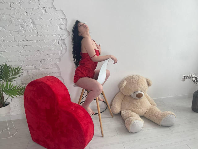 LiyaFrench with black hair, red lace dress, sitting on white chair, back against white wall, head tilted up, next to giant teddy bear and red heart-shaped chair.