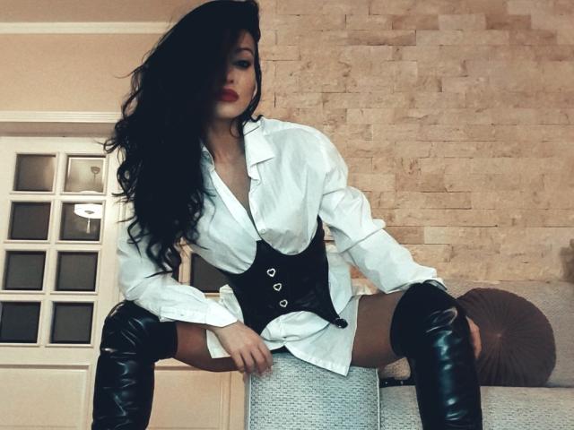 HotBisous with long black hair, red lipstick, white button-up shirt, black corset, and black thigh-high boots, sitting on a couch, showing her ass and pussy.