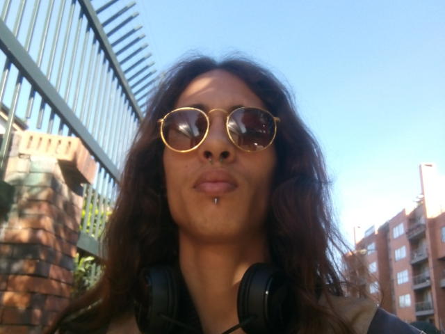 CalibanGray has a dark brown face, wavy hair, gold-rimmed sunglasses, a nose piercing, and black headphones. She's standing outside with a blue sky in the background.
