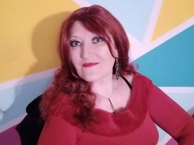 Sabrhina, avec ses cheveux roux, portant une robe rouge, un soutien-gorge noir et des boucles d'oreilles pendantes, a une poitrine de taille moyenne et un léger sourire, du rouge à lèvres rouge et une peau claire..