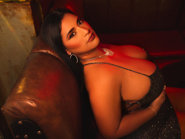 KorinaKova with large tits, wearing a sparkly black dress, leaning on a brown leather couch, dark hair, red lighting, hoop earrings, and a necklace, looking seductively at the camera.