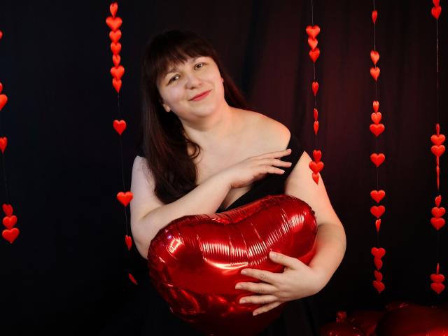 AliceInks holding a shiny red heart-shaped balloon, smiling, with red heart decorations hanging in the background, wearing a black and white off-shoulder top, fair skin, brown hair.