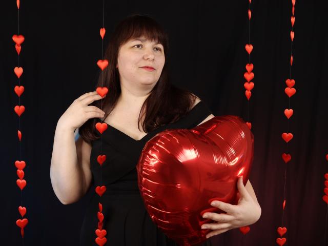 Alice, con capelli castani, vestito nero, che tiene un palloncino rosso a forma di cuore, con cuori rossi appesi sullo sfondo, con un leggero sorriso, la mano destra che tocca i capelli..