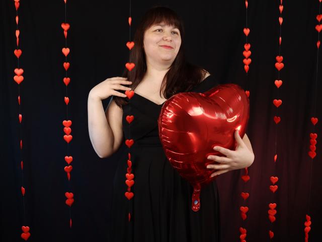 AliceInks with brown hair, black dress, holding shiny red heart-shaped balloon, smiling, standing against dark background with hanging red heart ornaments. Boobs are not visible.