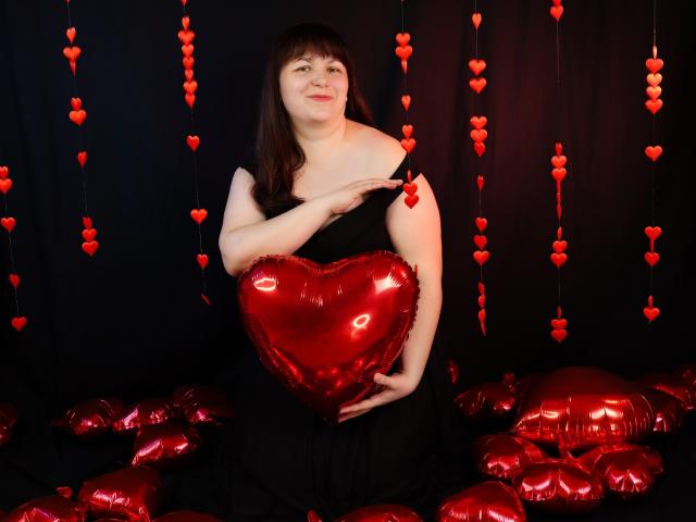AliceInks with pale skin, brown hair, and small tits, holding a shiny red heart-shaped booby, wearing a black dress, surrounded by red heart balloons, smiling.
