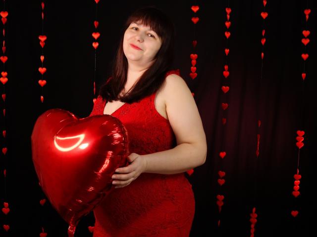 AliceInks with brown hair, red lace dress, holding shiny red heart-shaped balloon, smiling slightly, standing against black backdrop with red heart strings. Her tits are visible under dress.