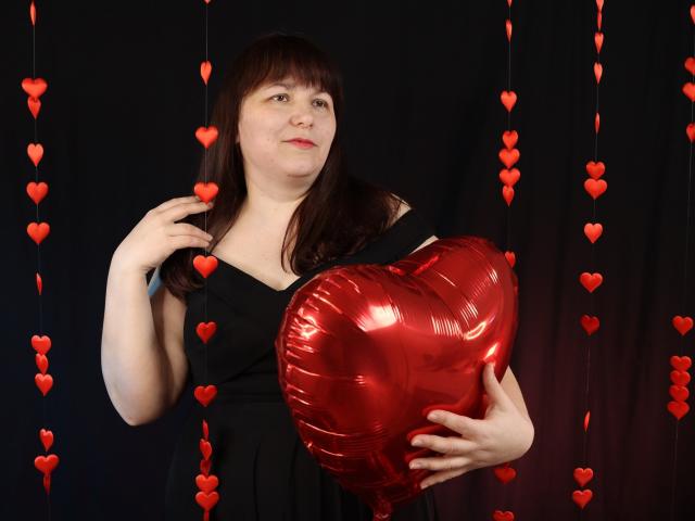 AliceInks, a chubby white woman with brown hair, holds a shiny red heart-shaped balloon in front of her, wearing a black dress, with red heart decorations hanging.