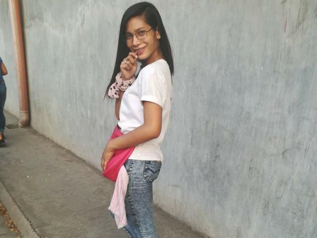 TrixieMattel with long black hair, glasses, white t-shirt, blue jeans, pink purse, holding pink and white clothes, smiling, standing against a gray textured wall, finger to mouth.