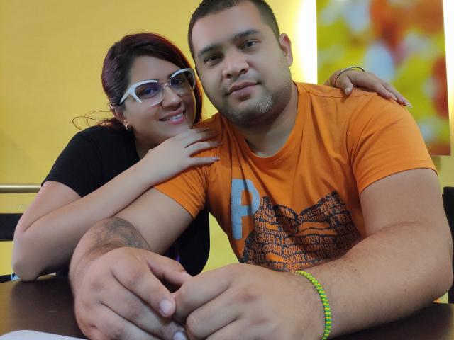 StevenAndPaty, with glasses and red hair, leans on a muscular guy in an orange shirt with a "P". He's wearing a neon bracelet, she's showing her tits.