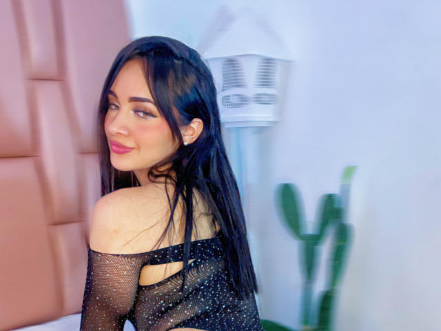 AngelCutie avec des cheveux noirs, portant un haut noir transparent, souriant à la caméra, montrant son épaule nue et le haut de son dos, assise près d'une tête de lit rose rembourrée..
