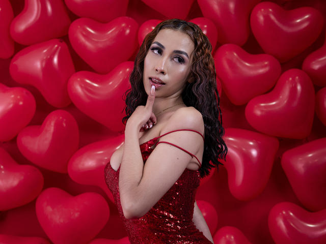 AnyaJay with wavy brown hair, wearing a red sequined dress, touching her finger to her lips, surrounded by red heart-shaped balloons. Her tits are visible, she's sexy.