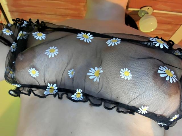 SarahGood's see-through black mesh bra with white daisy patterns covers her small tits, showing her nipples through the fabric. Her skin is pale. Ceiling light in background.