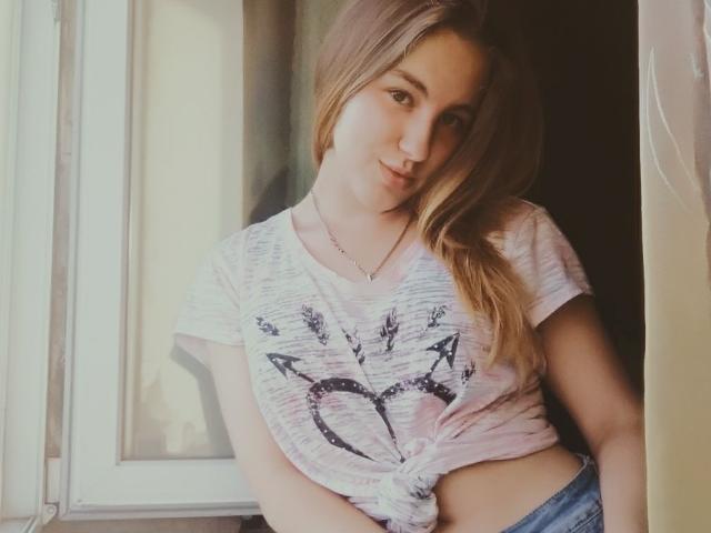 Una mujer con cabello rubio largo, camisa blanca atada que deja ver sus senos, estampado de corazón y flecha, apoyada en una ventana, sonriendo ligeramente, vestida con jeans azules, luz suave, pose relajada..