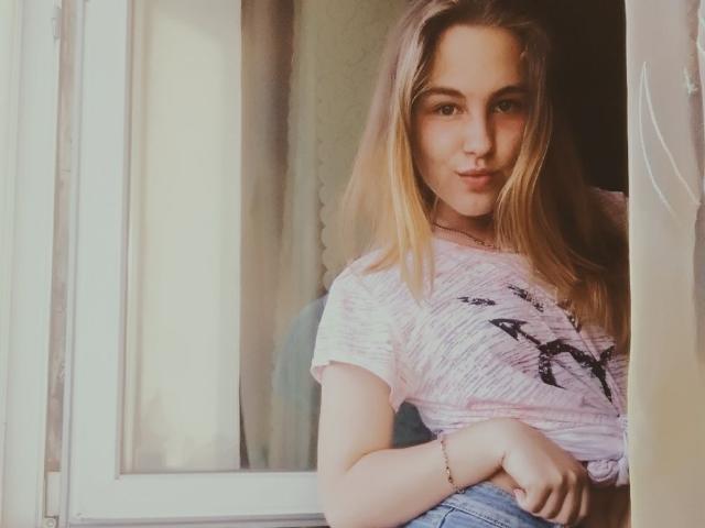 Dooris with blonde hair, pink shirt, and black design, leaning on window frame, smiling, hands on waist, wearing a bracelet, light skin, cute, soft lighting.