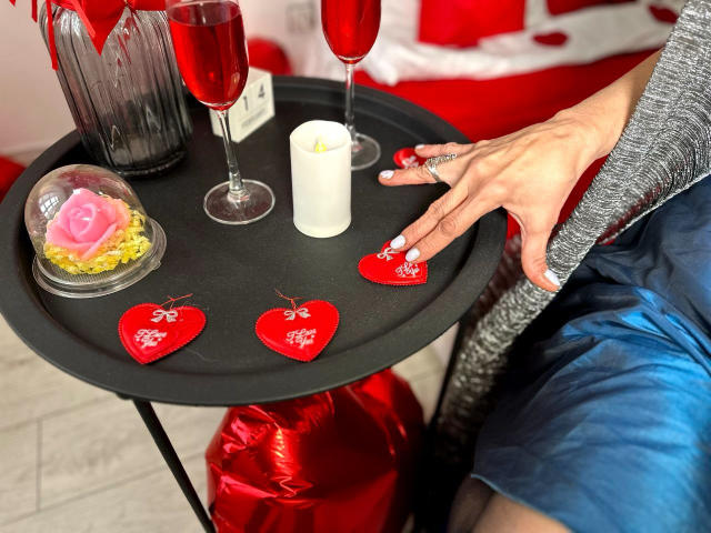 La main de SondraRiddle, portant une bague rouge, vêtue d'un pull gris et de shorts bleus, touche un plateau noir orné de cœurs rouges, de bougies et de verres à vin, tout en tenant une rose rose..