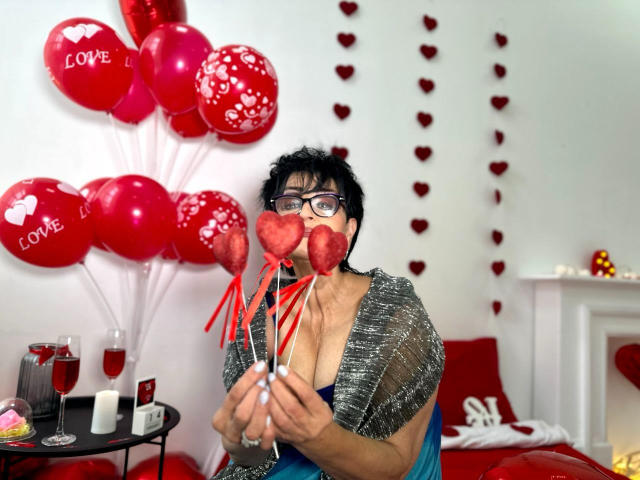 SondraRiddle with short black hair, glasses, and a gray shimmery top, holding red heart-shaped candies, surrounded by red balloons and heart decorations, sitting in a Valentine's-themed room.