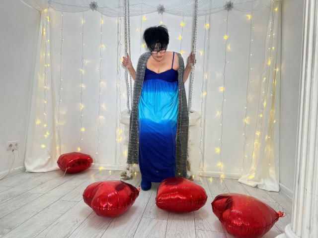 Sondra Riddle, con capelli neri, occhiali, vestito blu scuro, cardigan grigio, seduta su un'altalena bianca, circondata da cuscini a forma di cuore rossi, luci fatate gialle, in una stanza bianca..