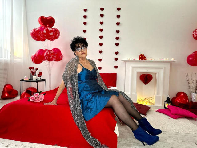 Sondra Riddle, com cabelo preto encaracolado, óculos, vestido azul, meias pretas, cardigan cinza e botas azuis, sentada em uma cama vermelha, cercada por corações e balões vermelhos..