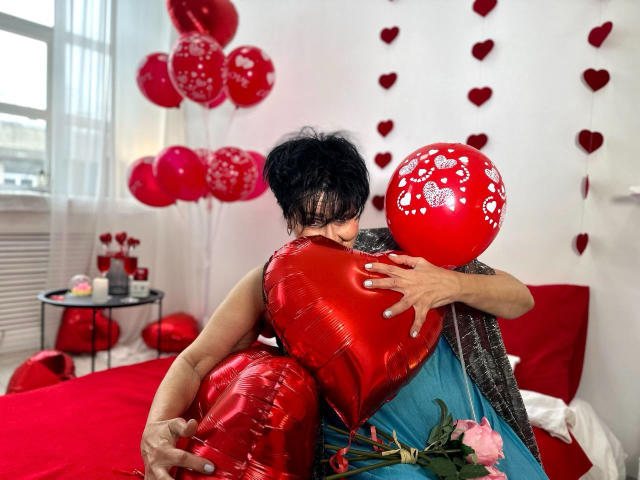 Sondra Riddle abbraccia un palloncino a forma di cuore rosso lucido, tenendone un altro con una pallina rossa sulla destra. Sullo sfondo ci sono palloncini rossi e decorazioni a forma di cuore..