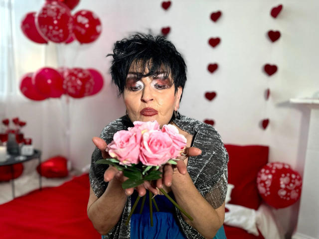 Sondra Riddle, avec ses courts cheveux noirs, tenant des roses roses, portant un pull gris en tricot et une robe bleue, regardant l'appareil photo avec une expression boudeuse..