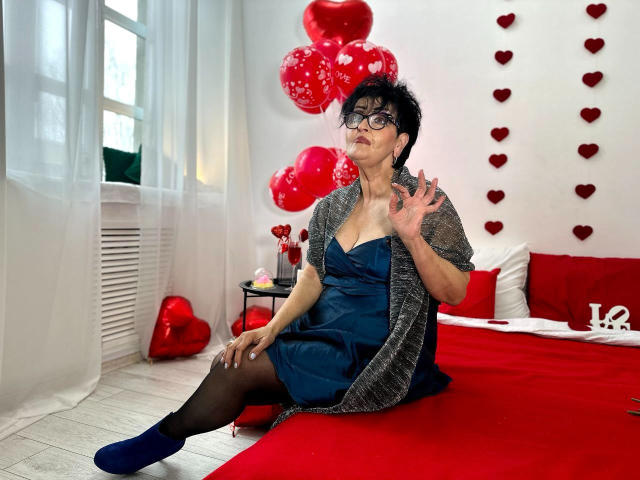 SondraRiddle with short black hair, glasses, blue dress, black stockings, grey cardigan, sitting on red bed, hand up, red balloons and hearts, white curtains, blue socks.