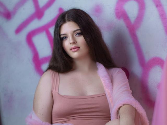 PamDares with long brown hair, pink tank top, and fluffy pink cardigan, looking cute with subtle makeup, showing small tits against a graffiti wall.