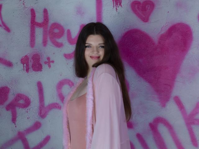 PamDares, avec de longs cheveux bruns, une robe rose et un cardigan en fausse fourrure rose, sourit à l'appareil photo, montrant légèrement ses seins sous sa robe, debout devant un mur rose recouvert de graffitis..