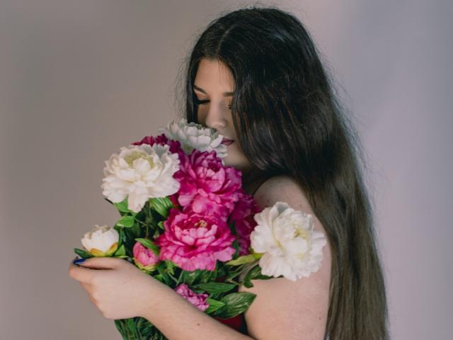 PamDares with long brown hair, holding a bouquet of pink and white peonies, eyes closed, smiling softly. Her tits are slightly visible, arms around the flowers.