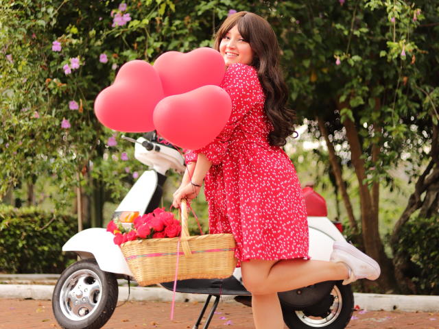 Tracie Lowe con un grupo de corazones rojos en forma de globo, vestido floral rojo, zapatillas blancas, cesta de mimbre con rosas, arrodillada sobre un patinete blanco, cabello ondulado castaño, sonriendo, exuberante jardín verde..