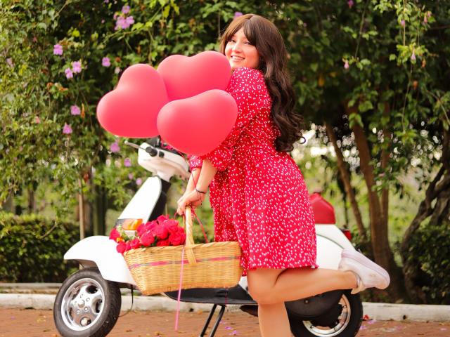 Tracie Lowe, mit langen braunen Haaren, in einem roten Kleid mit Blumenmuster, hält rosa Herzballons und kniet neben einem weißen Roller mit einem Korbkörbchen voller roter Blumen..