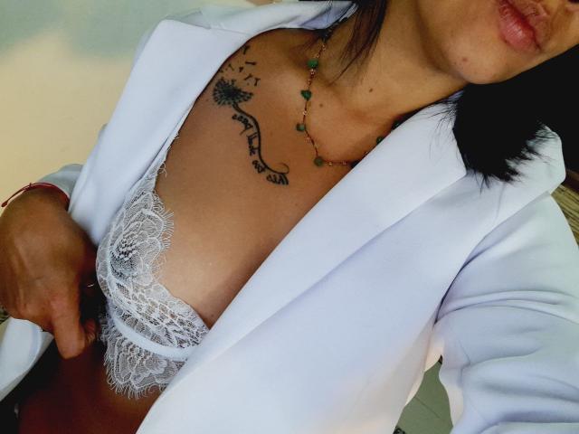 sashasweetx with black hair, wearing white blazer, lace bra, and green bead necklace, exposing tits, has tattoo on chest, right hand on blazer.