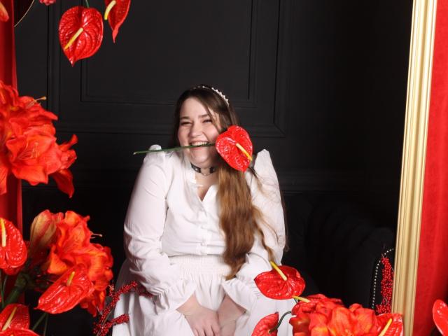 DaniellaGraceZZ with long brown hair, white dress, and choker, holding red lily in mouth, surrounded by red flowers, smiling, black wall, gold frame, red curtain.