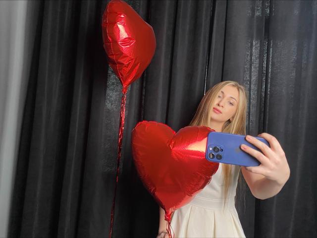 BeataAngel com cabelos loiros longos, vestindo um vestido branco, segurando um telefone azul, tirando uma selfie com balões vermelhos em formato de coração ao fundo..