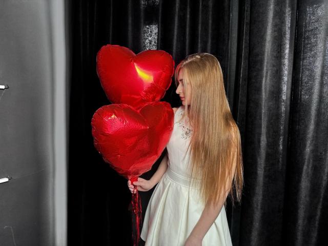 BeataAngel with long blonde hair, wearing a white dress, holding two shiny red heart balloons, standing in front of black curtains.