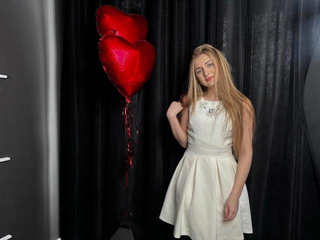 BeataAngel with long blonde hair, wearing a white dress, holding a red heart balloon, standing next to black curtains, smiling slightly, right hand touching her hair.