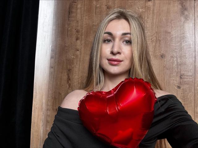 BeataAngel com cabelos loiros longos, blusa preta com um ombro à mostra, segurando um balão vermelho brilhante em formato de coração, sorrindo suavemente, pele clara, batom rosa, fundo de madeira, cortina preta à esquerda..