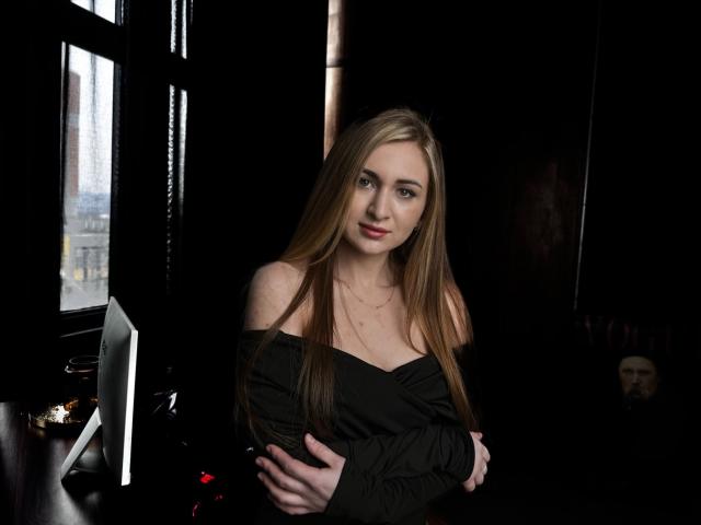 BeataAngel with long blonde hair, off-shoulder black dress, arms crossed, slight smile, standing in dark room, window to left, subtle light, looking at camera, small necklace.